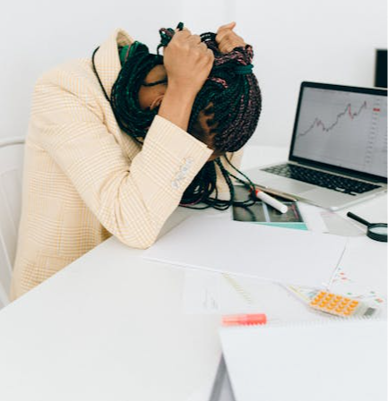 A stressed woman sits at her work desk with a laptop displaying stock market graphs.